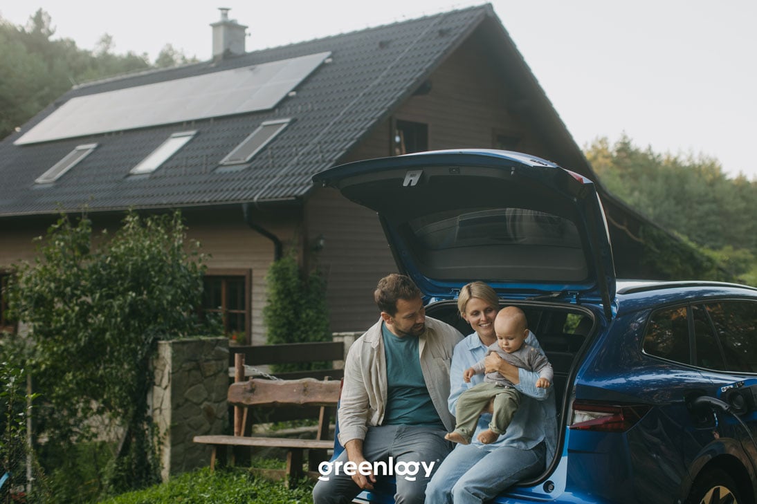 Šťastní rodičia s batoľaťom v náručí sediaci v otvorenom kufri auta pred domom s fotovoltickými panelmi na streche